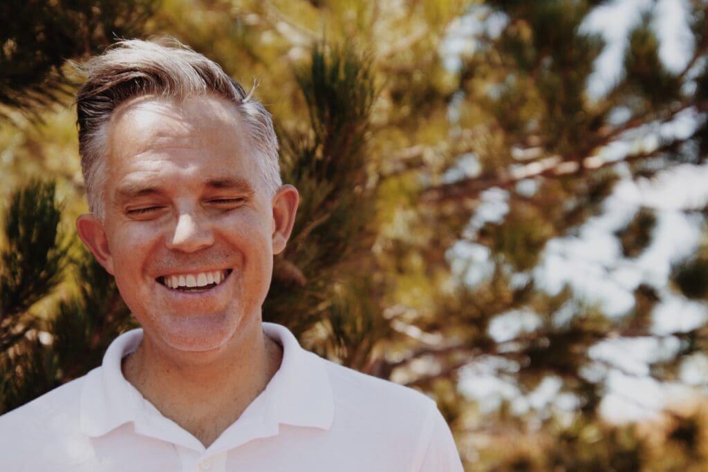 Man in white polo shirt smiling with green trees in background